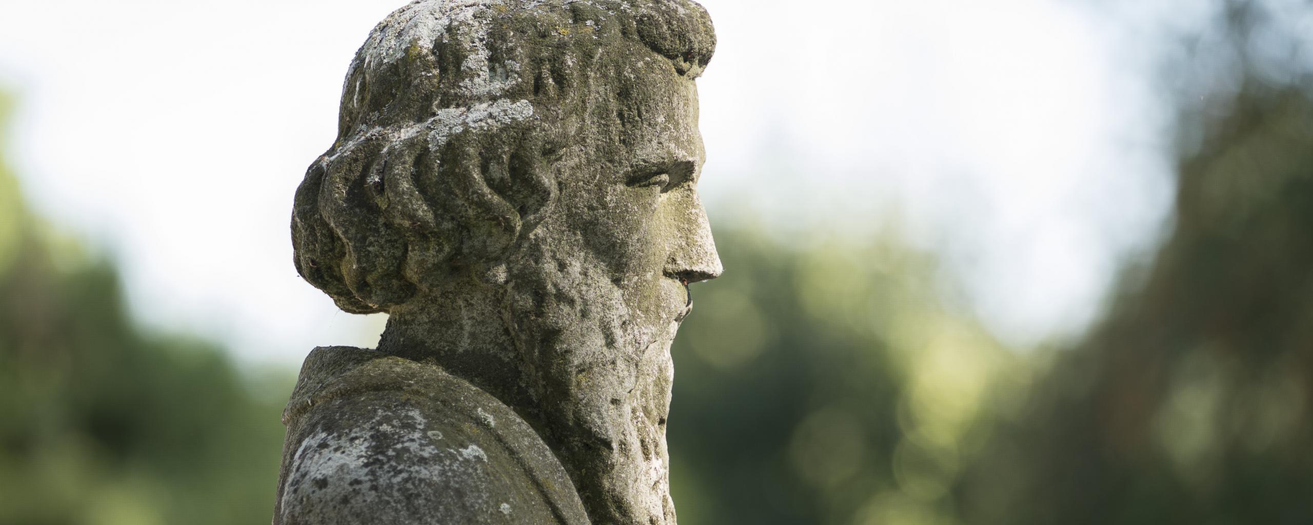 Een stenen beeld van een man met een baard die zijn handen vouwt. Op de achtergrond is het groen van bomen en struiken te zien. De zon schijnt op het gezicht van het beeld.