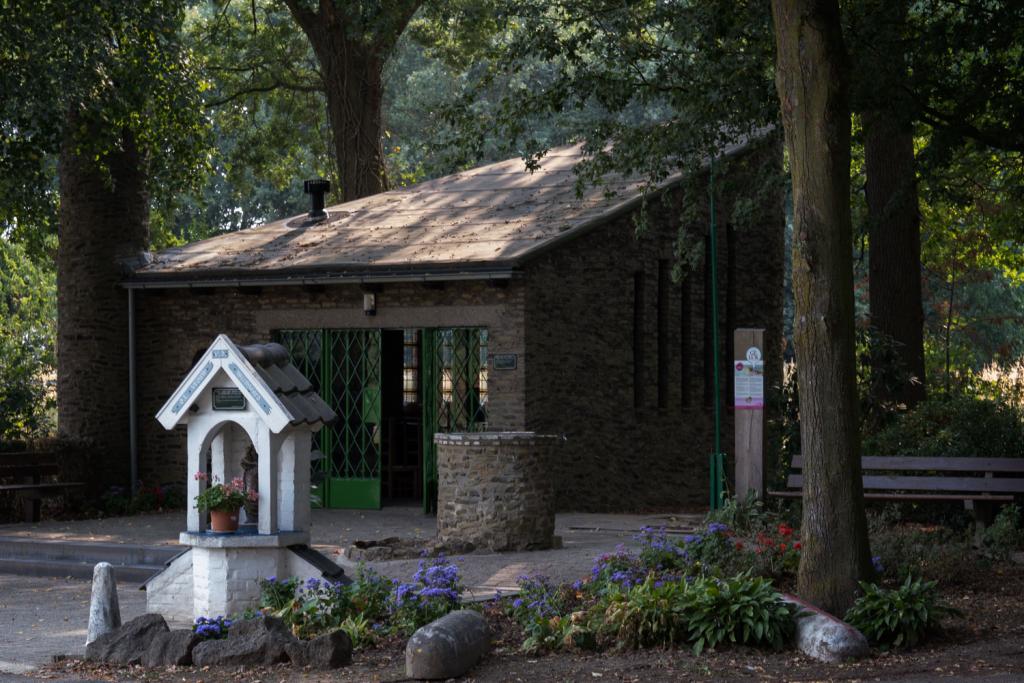 De twee kapellen die te vinden zijn aan de Wijngaardstraat. Links zien we het kleine aanbiddingskappelletje van witte stenen. Op de achtegrond de grote kapel van bruine stenen. Om de twee kapellen staan bomen en bloemen.