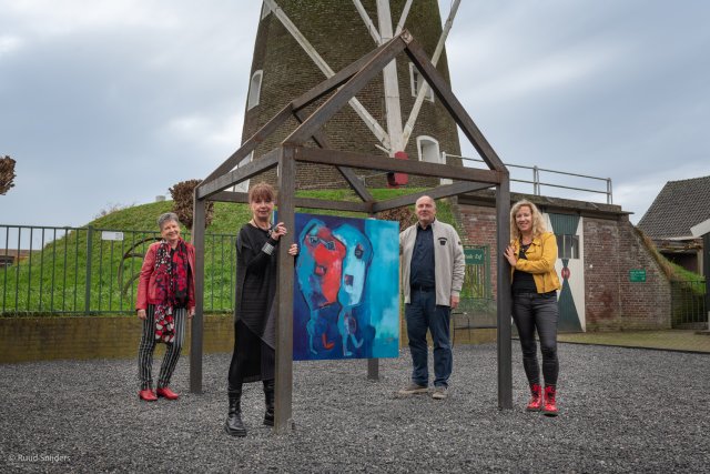 We zien de molen van Sint Odiliënberg op de achtergrond. Op de voorgrond een houden 'kunsthuisje' met daarin een schilderij. Op de foto staan vier kunstenaars om het kunsthuisje.