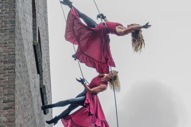 We zien twee vrouwen met rode jurken aan die aan kabels hangen. Ze zetten zich af tegen een kerktoren. Dit is een foto van een dansoptreden tijdens het Limburg Festival van 2021
