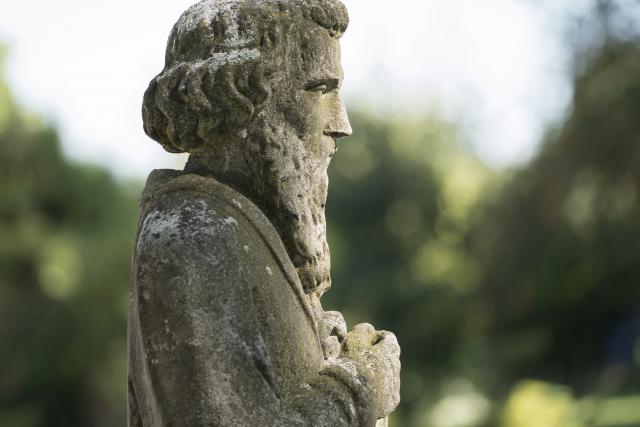 Een stenen beeld van een man met een baard die zijn handen vouwt. Op de achtergrond is het groen van bomen en struiken te zien. De zon schijnt op het gezicht van het beeld.