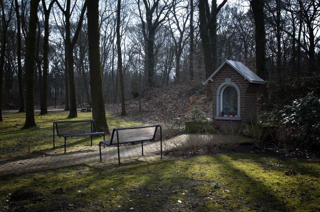 We zien een kapelletje en twee bankjes. Er loopt een pad naar het kapelletje toe, tussen de twee bankjes door. Achter de kapel begint een heuvel. De omgeving is omringt door bomen.