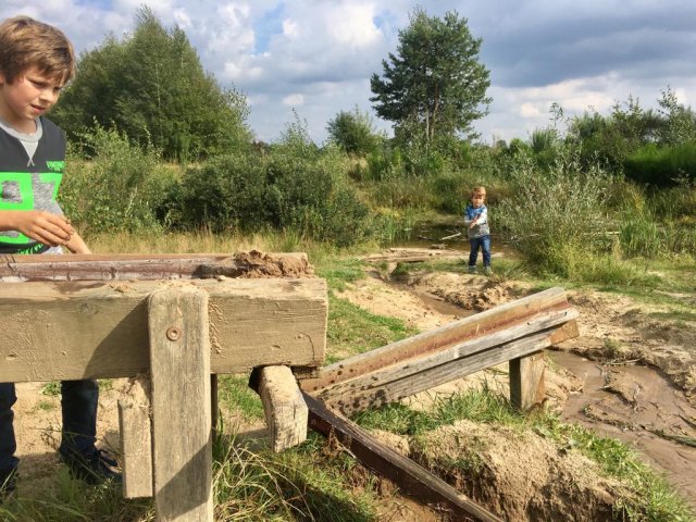 We zien twee kinderen spelen met houten verbindingen waar water doorheen stroomt. Het is de natuurspeelplaats bij Nationaal Park de Meinweg.