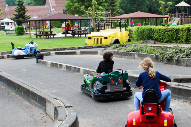 Twee auto's die op de racebaan rijden in speeltuin de Gitstap. Op de achtergrond zien we groen, een picknicktafel en een stenen gebouw.