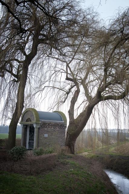 We zien de zijkant van een stenen kapelletje met rond, gebogen dak. De kapel staat op een heuvel en onderaan de heuvel loopt een beek. Boven de kapel zien we de kale takken van twee grote treurwilgen. Ze hangen over de kapel en over het water.