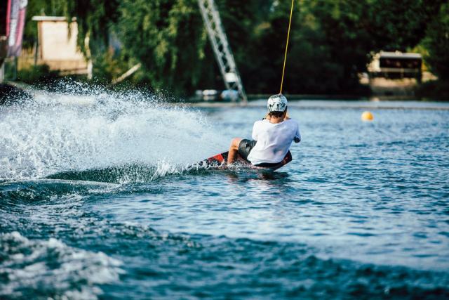 We zien een man die aan het waterboarden is op het meer van Amici Beach. Hij houdt zich vast aan een touw en zijn voeten zitten vast aan een board. Op de achtergrond zien we een deel van het strand.